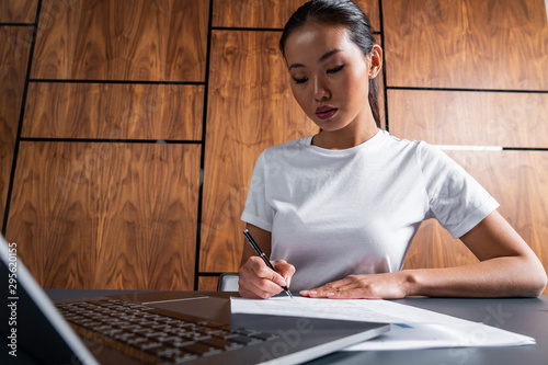 Serious young Asian businesswoman in casual clothes signing document at office table. Concept of employment, business and finance