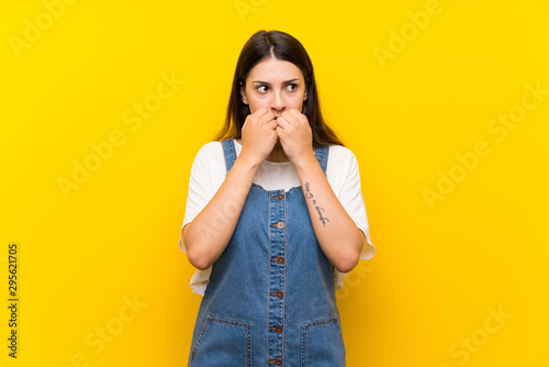 Young woman in dungarees over isolated yellow background nervous and scared putting hands to mouth