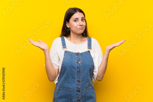 Fotografie Young woman in dungarees over isolated yellow background making doubts gesture