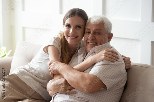 Adult granddaughter and elderly grandfather embracing sitting on couch indoors