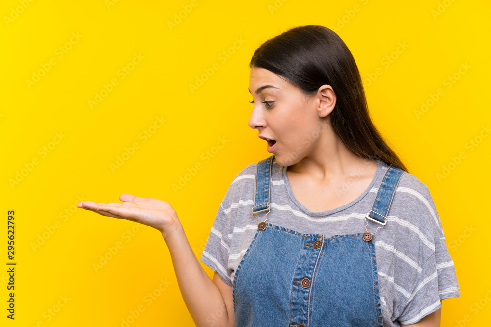 Young woman in dungarees over isolated yellow background holding copyspace imaginary on the palm Young woman in dungarees over isolated yellow background holding copyspace imaginary on the palm