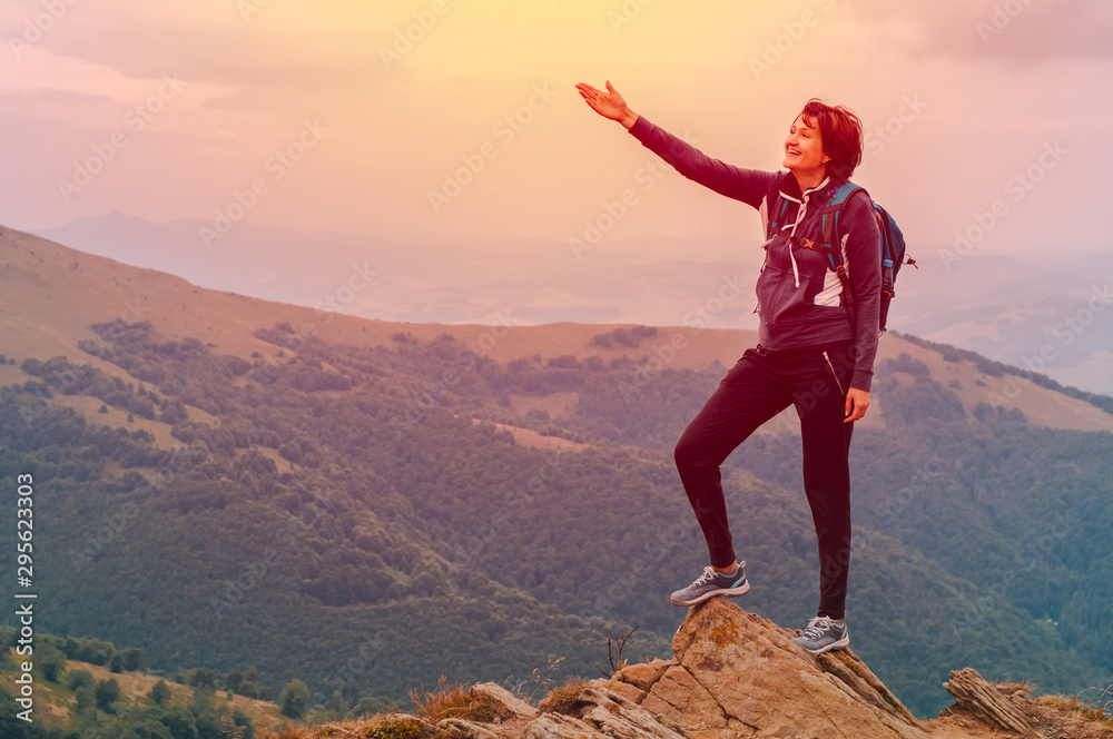Fototapeta premium Young girl tourist with backpack behind her stands on top of the mountain and enjoys a view of the Carpathian mountains at sunrise.
