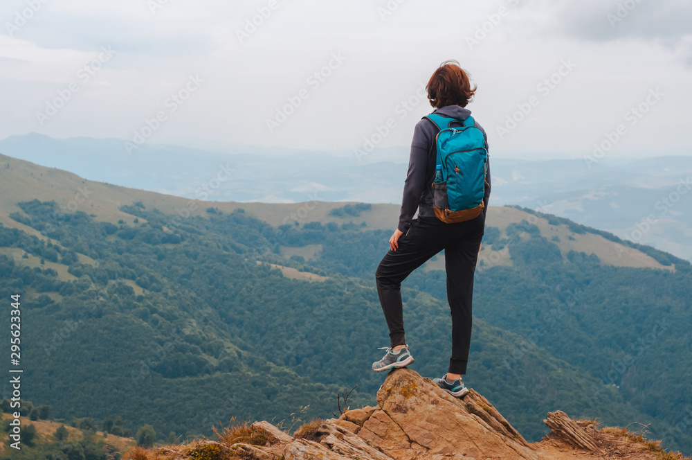 Naklejka premium Young girl tourist with backpack behind her stands on top of the mountain and enjoys a view of the Carpathian mountains at sunrise.
