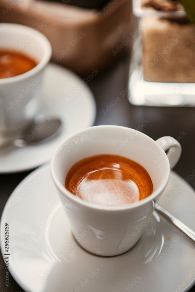 Close up. The small white ceramic mug (cup) with Italian espresso (coffee) on a table with a saucer and a metal spoon in a local Italian bar (coffee shop) in Milan, Lombardy, Italy. Beverage. Close up. The small white ceramic mug (cup) with Italian espresso (coffee) on a table with a saucer and a metal spoon in a local Italian bar (coffee shop) in Milan, Lombardy, Italy. Beverage.