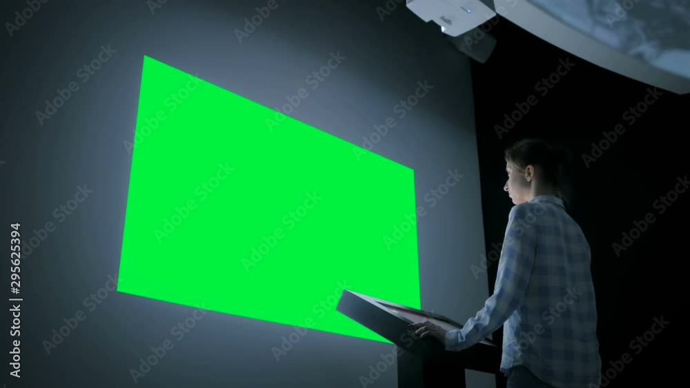 Mock up, futuristic, chroma key, template, green screen concept. Woman using electronic kiosk and looking at green blank large interactive wall display in dark room of modern technology museum