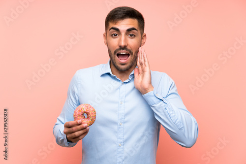 Young man holding a donut over isolated pink background with surprise and shocked facial expression