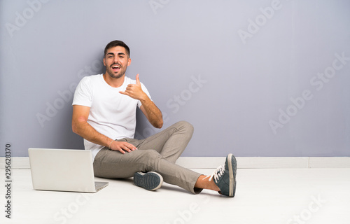 Young man with his laptop sitting one the floor making phone gesture