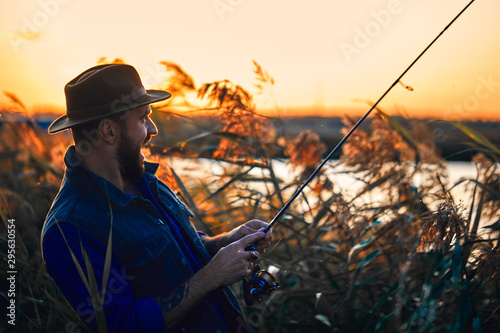 Caucasian bearded man in hat, smile fishing in reed near lake. Sunset. photo