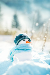 Little snowman made of snow in a knitted hat and a blue scarf on a background of a winter snowy landscape on a sunny frosty day.