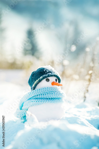 Little snowman made of snow in a knitted hat and a blue scarf on a background of a winter snowy landscape on a sunny frosty day. © isavira