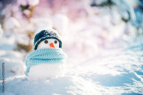 Little snowman made of snow in a knitted hat and a blue scarf on a background of a winter snowy landscape on a sunny frosty day. © isavira