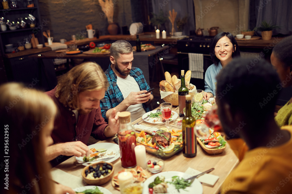 Multi-ethnic group of friends enjoying delicious dinner together sitting at table in dimly lit room, focus on smiling Asian woman