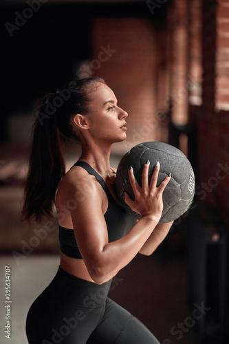 Slim flexible female in black sportswear do exercises in fitness gym. Brickwall background