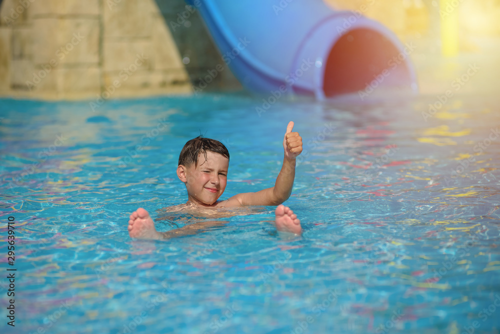 Active Caucasian boy spending his summer holidays in water park, he splashing in pool against colorful plastic slide.