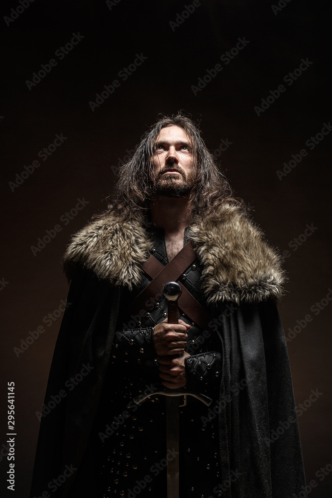 Man dressed in medieval armor and raincoat with longs word fighting against enemy. Courage fantasy warrior knight with long hair concept historical photo - obrazy, fototapety, plakaty 