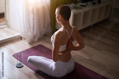 Caucasian short-haired woman practicing yoga with namaste behind the back, sitting in seiza exercise, vajrasana pose. Wearing in white leggins and white topic. Meditating towards window
