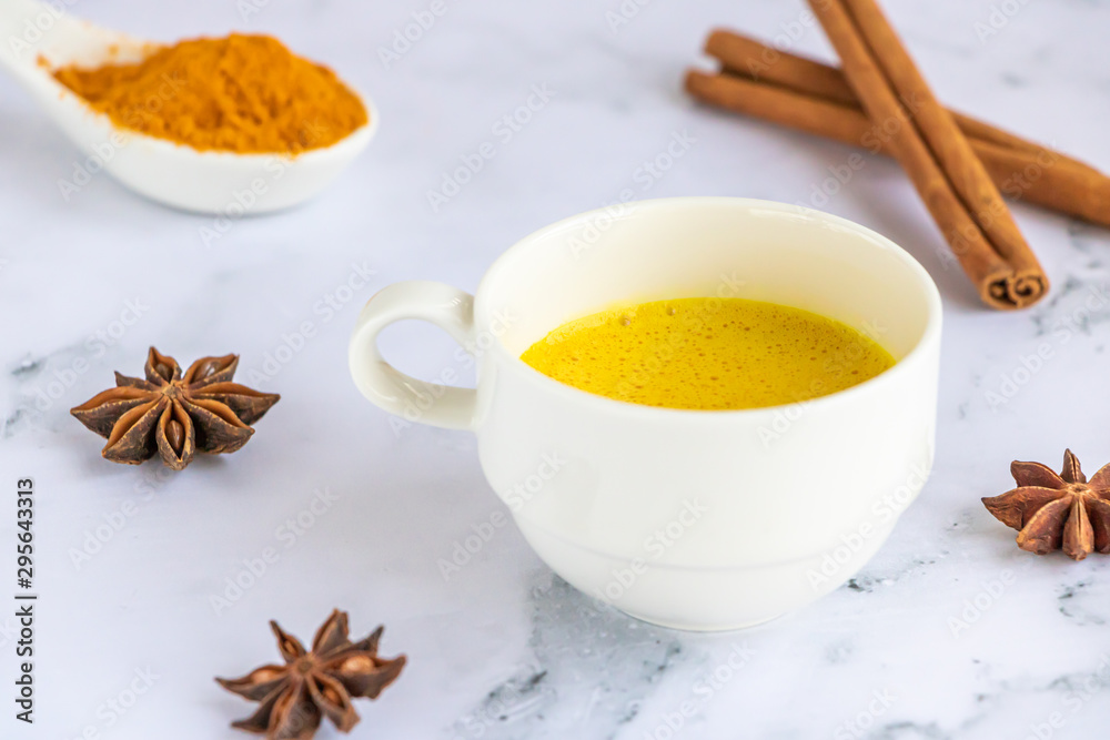 Golden (turmeric) milk in a white cup on the marble background. A spoon with turmeric powder. Cinnamon and anise. Golden (turmeric) milk in a white cup on the marble background. A spoon with turmeric powder. Cinnamon and anise.