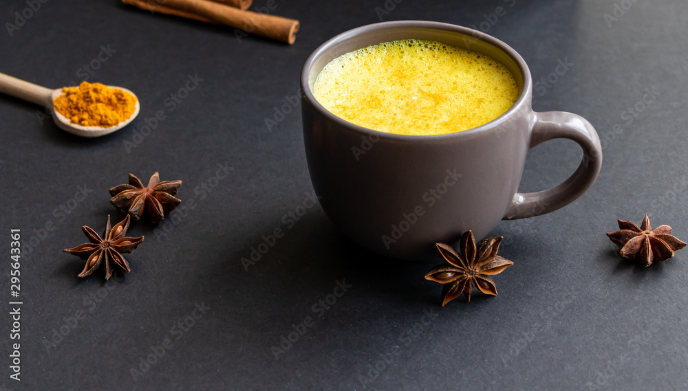 Golden (turmeric) milk in a gray cup on the dark background. A spoon with turmeric powder. Cinnamon and anise. Golden (turmeric) milk in a gray cup on the dark background. A spoon with turmeric powder. Cinnamon and anise.