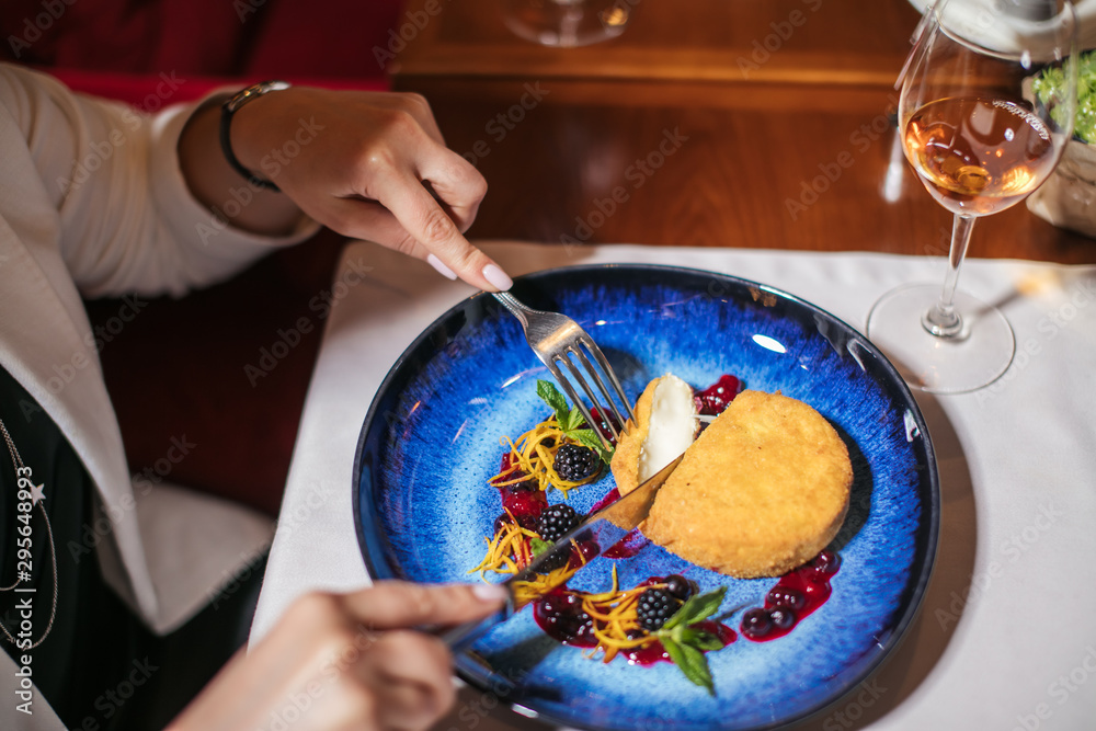 Obraz premium Specially prepared sweet dessert served at table on original beautiful blue plate, prettified with berries and green leafs of mint, gentle hands cuts a small piece of soft cake with silver knife