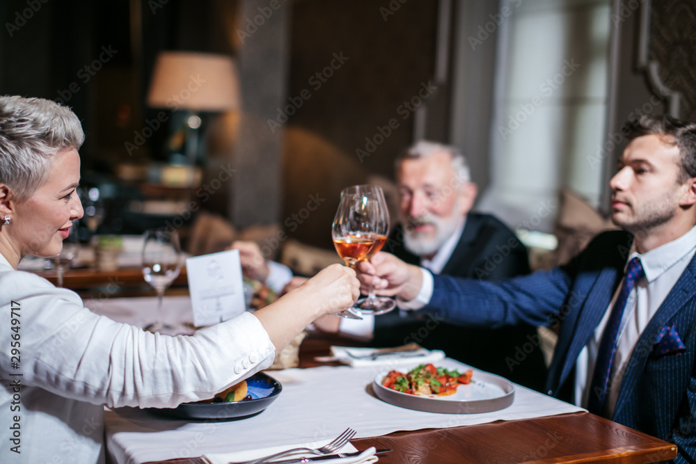 Responsible and hard working colleagues celebrate successful deal consummation , drink fragrant wine, clink glasses, smile with cheerful expression in restaurant, horizontal shot