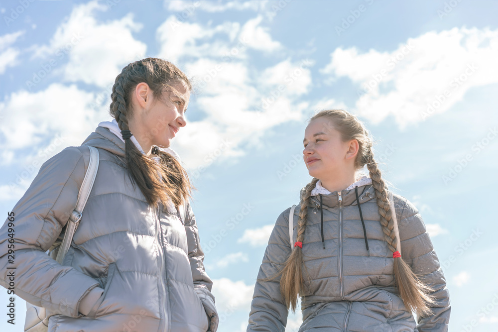 two girlfriends teenagers with pigtails in warm gray jackets walk on street blue sky with clouds background