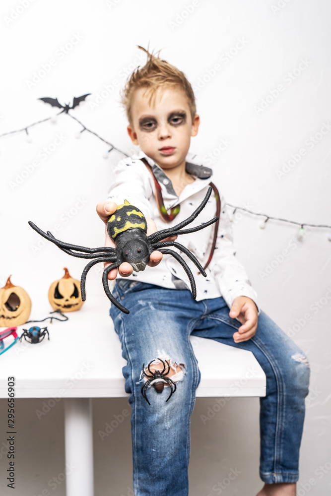 A little boy in a zombie costume holds a large spider in his hand.