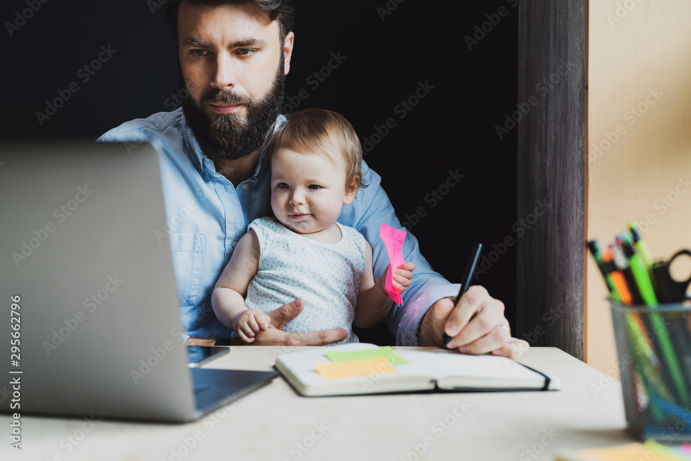 Parent with child studying remotely at homebased laptop. Distant education for people with babies.Man and toddler sitting at table Parent with child studying remotely at homebased laptop. Distant education for people with babies.Man and toddler sitting at table