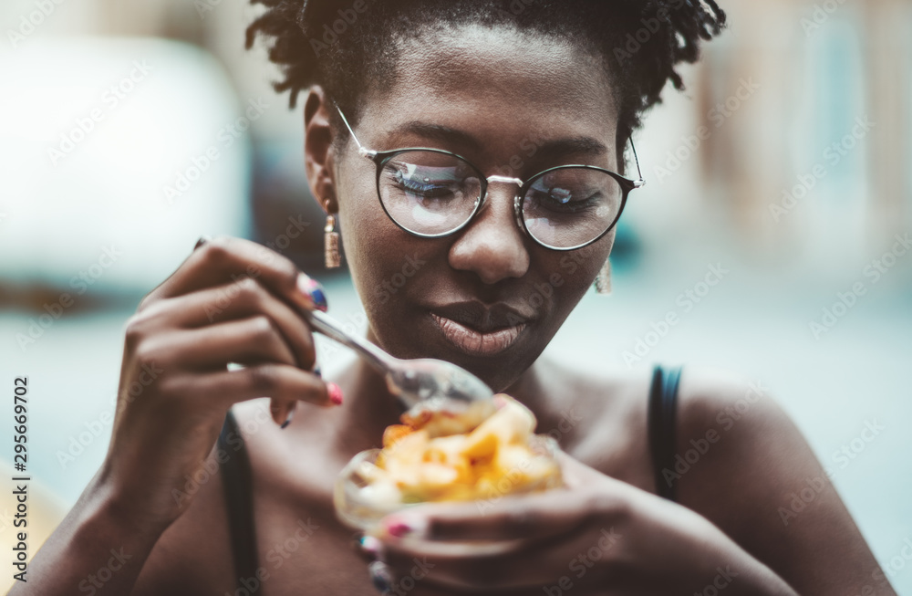 A portrait of a cheerful black girl in eyeglasses eating a delicious dessert in an outdoor cafe, selective focus, shallow depth of field; young smiling African female is eating ice cream in street bar A portrait of a cheerful black girl in eyeglasses eating a delicious dessert in an outdoor cafe, selective focus, shallow depth of field; young smiling African female is eating ice cream in street bar