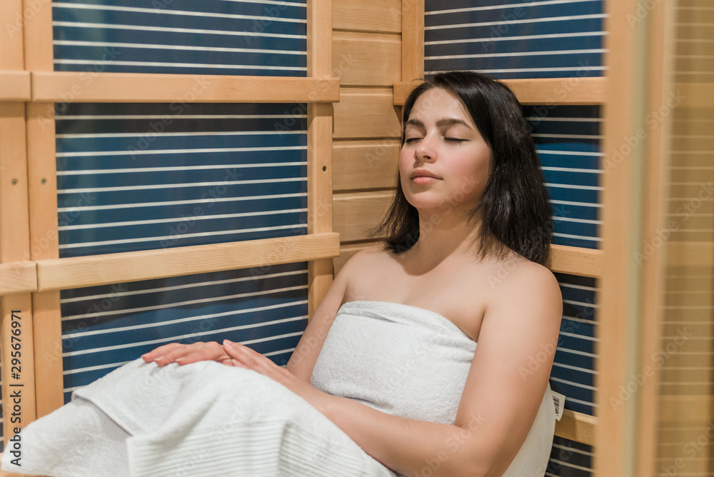 Young woman relaxing in a sauna dressed in a towel. Interior of new Finnish sauna, infrared panels for medical procedures, classic wooden sauna. Young woman relaxing in a sauna dressed in a towel. Interior of new Finnish sauna, infrared panels for medical procedures, classic wooden sauna.