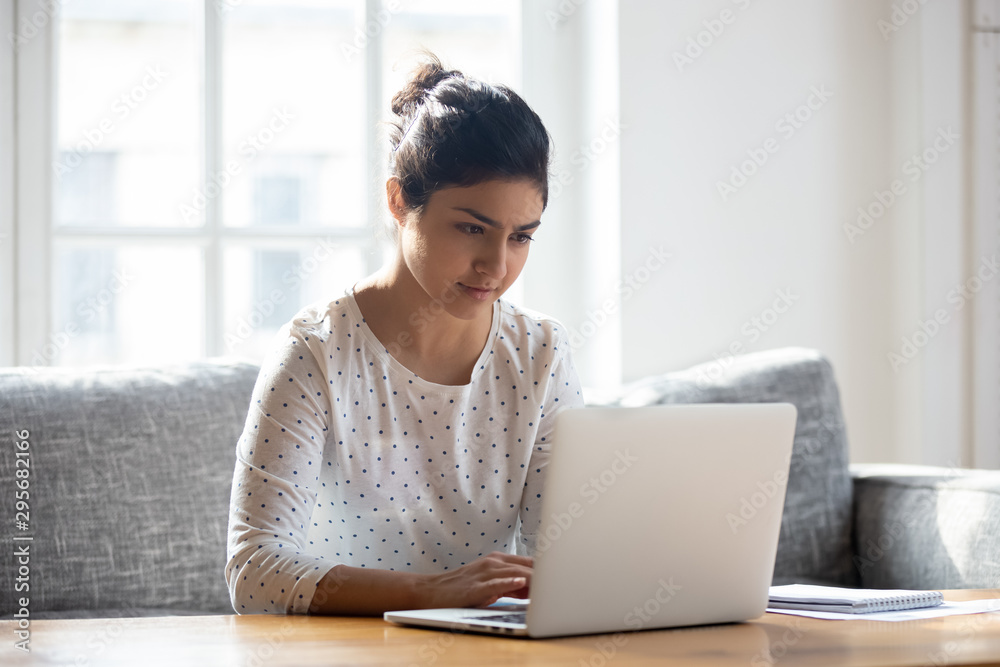 Focused Indian woman using laptop at home, looking at screen Focused Indian woman using laptop at home, looking at screen