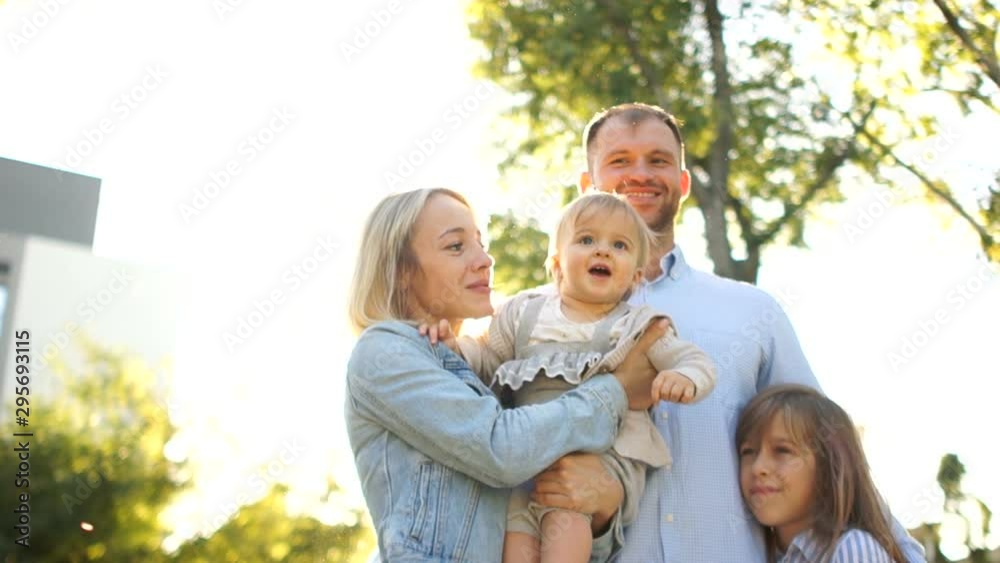 Family picnic in the park. Parents and their two daughters are hugging on the background of green foliage and bright sky Family picnic in the park. Parents and their two daughters are hugging on the background of green foliage and bright sky