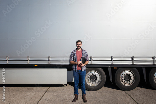 Portrait of middle aged bearded trucker standing in front of truck trailer against grey shiny tarpaulin. Copy space around truck driver. Transportation services.