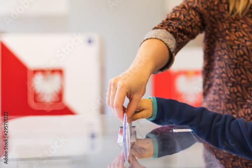 Woman's and kid's hands puting a card with a vote.to the ballot box during parliamentary elections. In the backround polish arms and flag photo