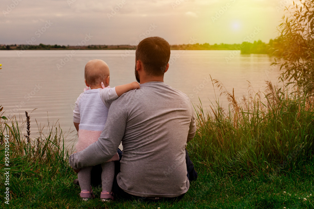 Fototapeta premium Daughter with father on the shore at sunset. Autumn, view from the back. Fatherhood and care.