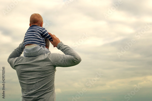 A child on his father’s neck. Walk near the water. Baby and dad against the sky.