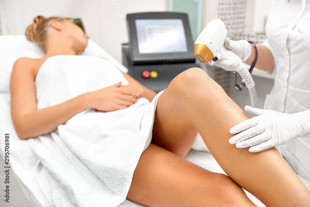 Fototapeta premium The girl is lying on the couch in the medical glasses in the treatment room. The hands of the cosmetologist make the epilation of the legs