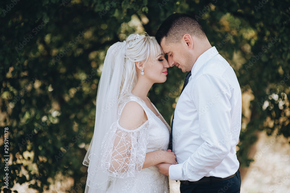 Beautiful newlyweds hug on the background of nature and trees. Wedding closeup portrait of a smiling groom and a cute blonde bride in a white dress. Photography and concept.
