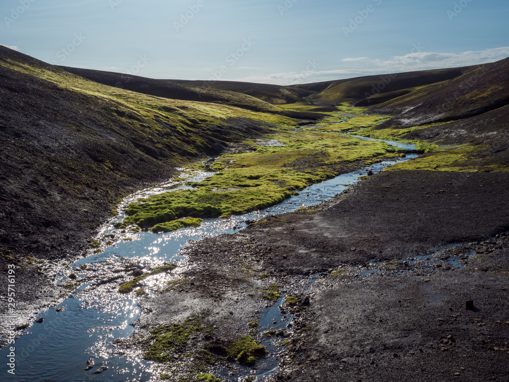 Small stream from melting ice with Lush vivid green wet moss in black lava hills in Landmannalaugar mountain. Fjallabak Nature Reserve in Highlands of Iceland, summer blue sky. Small stream from melting ice with Lush vivid green wet moss in black lava hills in Landmannalaugar mountain. Fjallabak Nature Reserve in Highlands of Iceland, summer blue sky.
