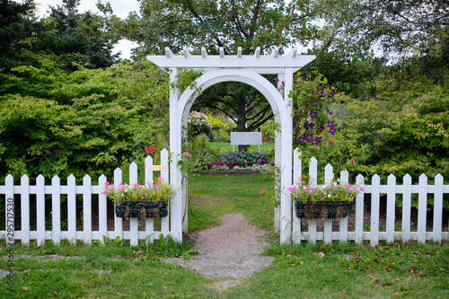 A white wooden arbour erected over a well worn foot path to a flower and shrub garden. The white latticework is covered in vines. Two boxes of pink flowers hangs from a white picket fence. 