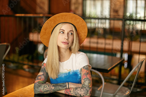 Indoor photo of attractive tattooed blonde woman in white and blue t-shirt and wide hat sitting over cafe interior and keeping folded hands on table, looking aside with light smile