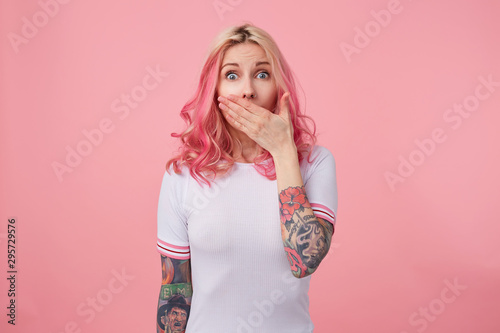 Indoor photo of open-eyed tattooed young female posing over pink background in casual clothes, looking to camera surprisedly and raising palm to cover opened mouth