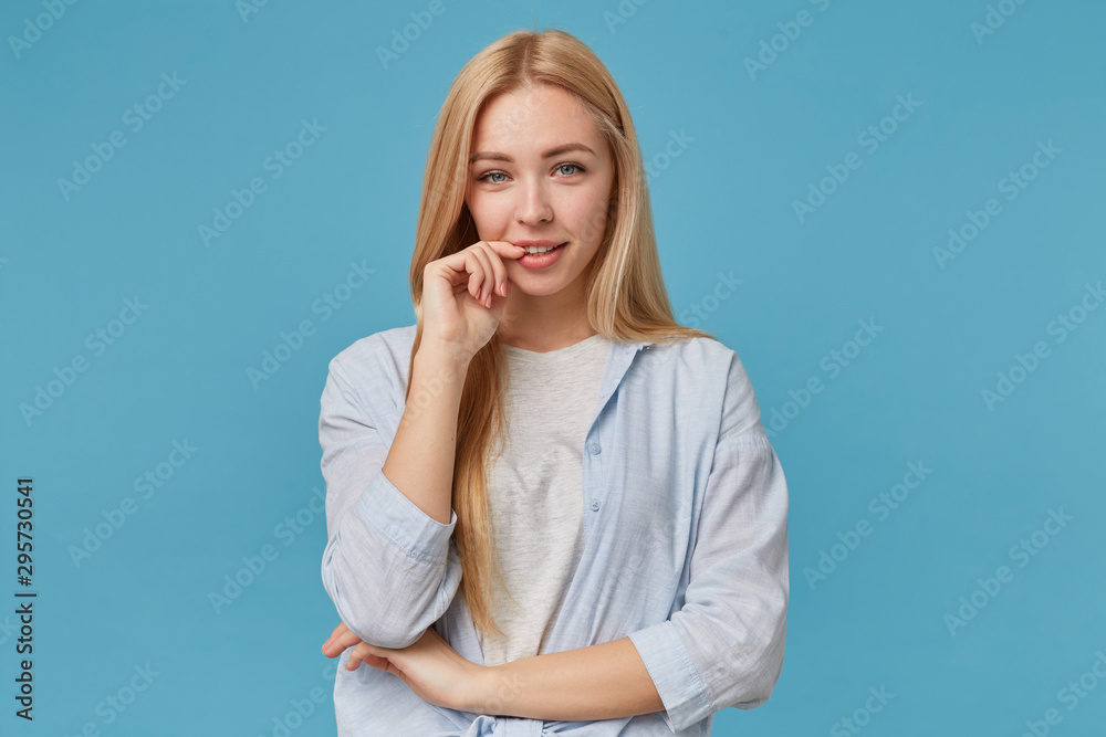 Fototapeta premium Horizontal photo of positive blonde long haired woman in casual clothes posing over blue background, keeping index finger on underlip and smiling to camera sincerely
