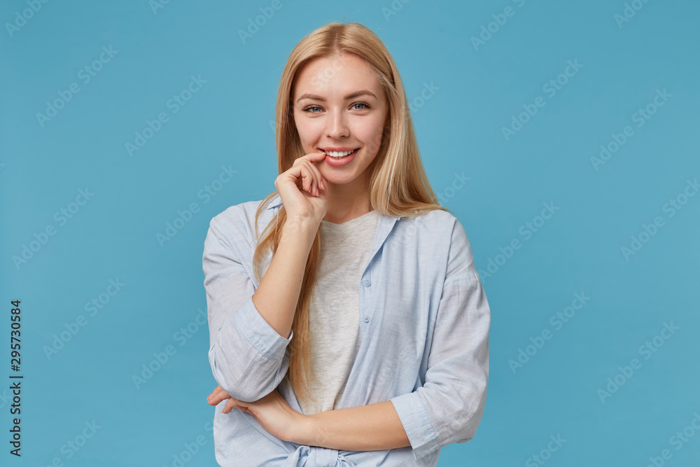 Smiling positive blonde female with attractive look and long hair posing over blue background, holding index finger on underlip and looking at camera cheerfully, wearing casual clothes Smiling positive blonde female with attractive look and long hair posing over blue background, holding index finger on underlip and looking at camera cheerfully, wearing casual clothes