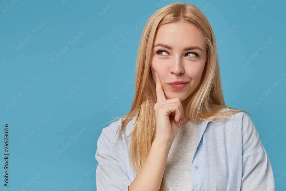 Close-up of beautiful young blonde lady with casual hairstyle looking aside thoughtfully, keeping forefinger on her cheek and smiling cunningly, isolated over blue background Close-up of beautiful young blonde lady with casual hairstyle looking aside thoughtfully, keeping forefinger on her cheek and smiling cunningly, isolated over blue background