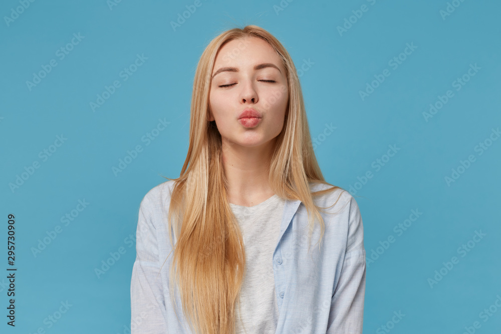 Studio photo of young lovely female with blonde long hair wearing casual clothes, posing over blue background with closed eyes and folding lips in air kiss Studio photo of young lovely female with blonde long hair wearing casual clothes, posing over blue background with closed eyes and folding lips in air kiss