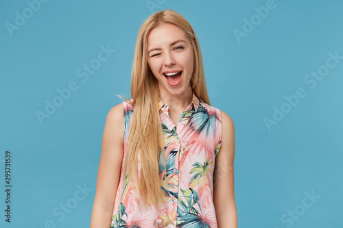 Cheerful attractive young blonde female with long hair wearing flowered shirt, standing over blue background with hands down, winking at camera with wide joyful smile