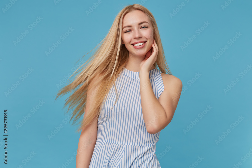 Portrait of attractive young blonde lady with long hair keeping hand on her face and smiling happily to camera, posing over blue background, demonstrating her white perfect teeth