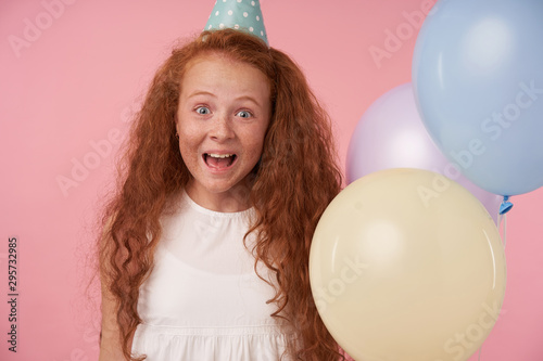 Portrait of joyous redhead girl with long curly hair in white dress and birthday cap being excited and surprised to get birthday present, happily looking in camera over pink background