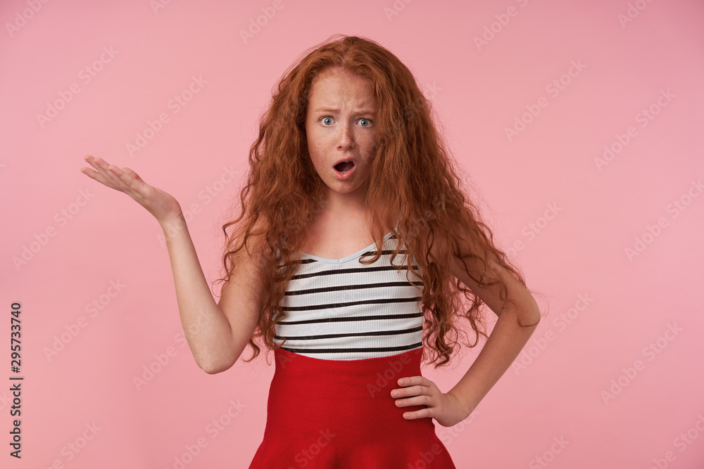 Portrait of redhead female kid with long curly hair posing over pink background in festive clothes, raising palm with confused face, looking at camera with wide mouth opened and frowning her face Portrait of redhead female kid with long curly hair posing over pink background in festive clothes, raising palm with confused face, looking at camera with wide mouth opened and frowning her face