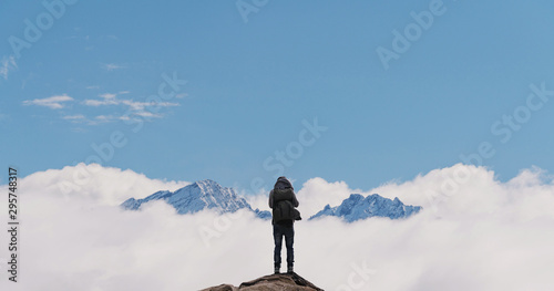 Canvas Print a man with backpack standing on mountain top, with snow mountains over clouds